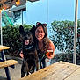 beer, bench, black_dog, canopy, casual, daylight, dog, glass, greenery, happy, leash, nature, outdoor, pet, picnic_table, portrait, smiling, string_lights, sunglasses, woman