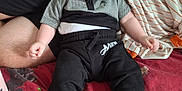 Roman joined the competition — help win amazing prizes! baby, infant, child, person, clothing, black_shoes, pants, shirt, bed, blanket, striped_blanket, red_blanket, sitting, indoors, cute, face, head, legs, feet, casual_wear