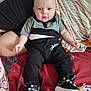 baby, infant, child, person, clothing, black_shoes, pants, shirt, bed, blanket, striped_blanket, red_blanket, sitting, indoors, cute, face, head, legs, feet, casual_wear