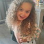 child, curly_hair, smiling, indoor, playful, jacket, shorts, heart_pattern, floor, table, bottles, cups, furniture, person, happy, young, pose, casual_clothing, room, light