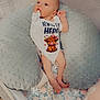 baby, infant, crib, onesie, cushion, floral_bedding, soft_toys, blanket, cute, newborn, clothing, sleeping_area, child, baby_bedding, indoors, person, footwear, mesh_sides, lying_down, alert