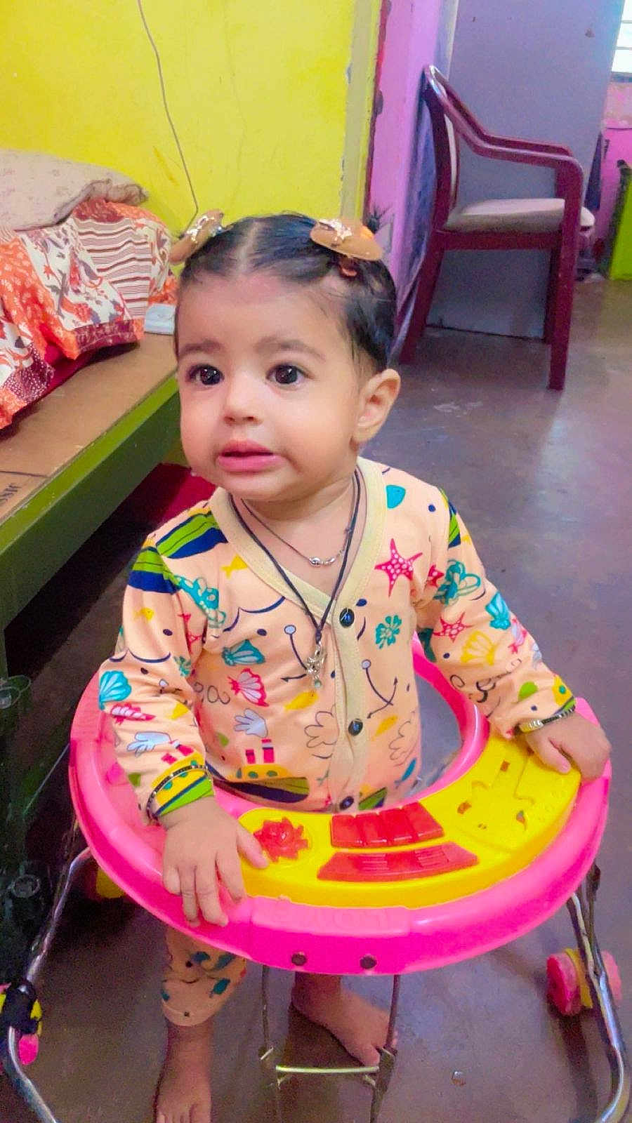 Avitaaz is registered to the contest to win money with this photo: toddler, walker, child, indoors, clothing, hair_clips, face, expression, footwear_barefoot, toy, bed, chair, floor, room, person, playful, colorful, curious, baby, jumpsuit