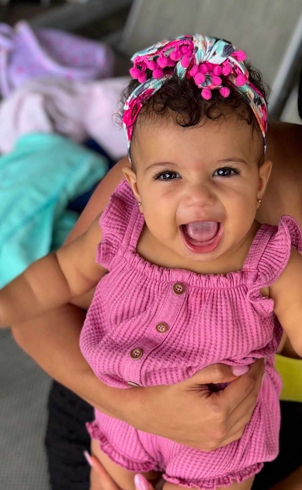 Daniella is registered to the contest to win money with this photo: baby, child, smiling, pink_outfit, headband, happy, person, cute, portrait, infant, hands, holding, curly_hair, earrings, buttoned_clothing, indoor, skin, fashion, closeup, expressive