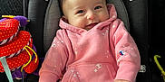 Lilia is registered to the contest to win money with this photo: baby, infant, smiling, car_seat, pink_suit, hoodie, plush_toy, colorful_toy, safety_label, seat_cushion, hands, face, eyes, hair, cozy, portrait, indoor, child, clothing, happy