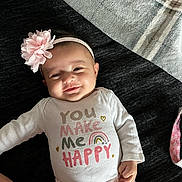 Lilia is registered to the contest to win money with this photo: baby, infant, smile, headband, flower_headband, onesie, text_on_clothing, rainbow_graphic, blanket, bedspread, hand, arm, face, cheek, pink, white_clothing, lying_down, happy, portrait, cute
