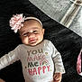 baby, infant, smile, headband, flower_headband, onesie, text_on_clothing, rainbow_graphic, blanket, bedspread, hand, arm, face, cheek, pink, white_clothing, lying_down, happy, portrait, cute