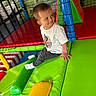 child, boy, playground, indoor_play_area, colorful, soft_play, netting, padded_surface, smiling, casual_clothing, white_shirt, dark_pants, socks, play_equipment, fun, happy, active, leisure, young, portrait