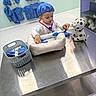 toddler, child, toy, stuffed_animal, dalmatian, doctor_costume, scrubs, stethoscope, medical_toys, play_clinic, blue, hat, basket, table, floor, shelf, indoor, pretend_play, cute, focused