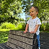 toddler, child, bench, outdoor, greenery, sunlight, white_tshirt, blue_jeans, happy, smiling, nature, wood, park, daylight, portrait, person, casual_clothing, expression, curious, standing