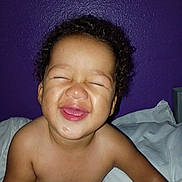 Willow is registered to the contest to win money with this photo: baby, infant, child, portrait, smiling, happy, eyes_closed, curly_hair, face, bare_shoulders, bed, bedding, pillow, bedroom, purple_wall, indoors, close_up, sitting, joyful, photography