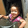 Willow is registered to the contest to win money with this photo: child, toddler, baby, smiling, portrait, stroller, seated, pink_shirt, curly_hair, brown_skin, face, eyes, outdoor, indoor, happy, harness, playful, messy_face, snack, photography
