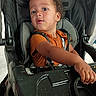 Willow joined the competition — help win amazing prizes! toddler, child, baby, stroller, seated, portrait, face, curly_hair, orange_shirt, harness, buckled, looking_at_camera, indoors, daylight, close_up, arm, hand, snack_crumbs, black_stroller, curious