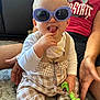 Renley is registered to the contest to win money with this photo: baby, sunglasses, tongue, hand, person, carpet, overalls, checkered, toy, teether, adult, sitting, smile, clothing, indoor, cute, infant, playful, support, comfort
