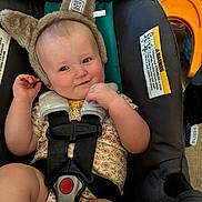 Renley is registered to the contest to win money with this photo: baby, child, car_seat, safety_harness, hat, bear_ears, smile, face, infant, cute, indoors, floral_outfit, straps, seat, toy, hand, skin, expression, comfort, padding