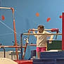 activity, balance, blue_wall, candid, child, concentration, equipment, exercise, fitness, gymnastics, gymnastics_bars, indoor, person, pink_shorts, red_padding, sport, stacked_mats, training, white_top, young