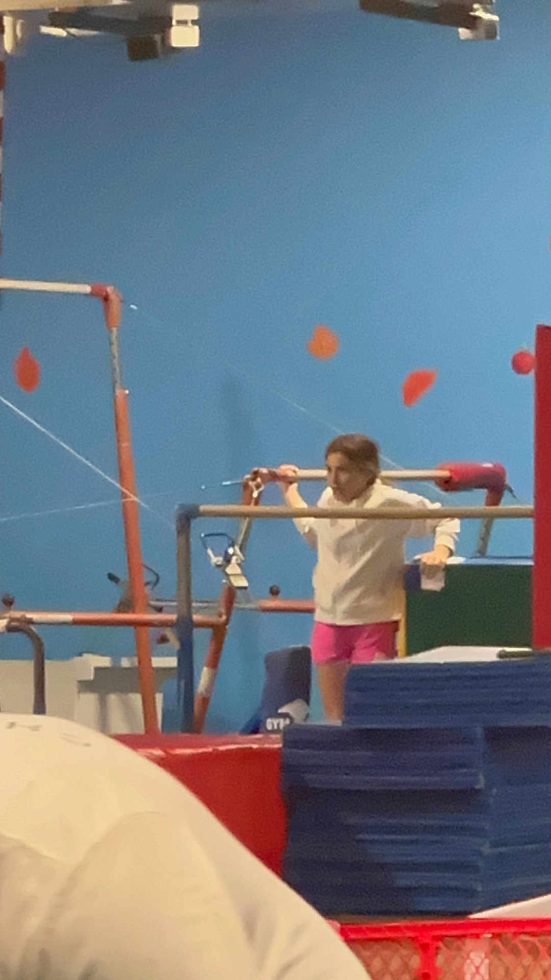 Izzy is registered to the contest to win money with this photo: child, gymnastics, gymnastics_bars, indoor, blue_wall, red_padding, stacked_mats, pink_shorts, white_top, exercise, sport, training, young, person, activity, equipment, fitness, balance, concentration, candid