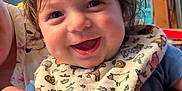 Wesley is registered to the contest to win money with this photo: baby, smile, bib, curly_hair, indoor, child, happy, person, window, hand, animal_print, clothing, face, portrait, young_child, holding, cute, skin, expression, colorful
