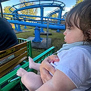 Wesley is registered to the contest to win money with this photo: child, toddler, baby, person, amusement_park, roller_coaster, ride, green_rail, blue_track, fence, trees, sky, outdoor, daylight, hair, hand, clothing, arm, legs, seat