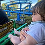 child, toddler, baby, person, amusement_park, roller_coaster, ride, green_rail, blue_track, fence, trees, sky, outdoor, daylight, hair, hand, clothing, arm, legs, seat