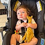 baby, child, car_seat, plush_toy, giraffe_toy, blue_eyes, safety_straps, white_onesie, curly_hair, indoors, floor, black_car_seat, infant, toy, cute, seated, expression, hand_holding, foot, stroller