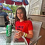 child, girl, smiling, red_shirt, sprite_can, glass_table, outdoor, umbrella, samuel_adams, restaurant, dining, chair, wristband, wooden_wall, people, summer, casual, happy, daylight, portrait