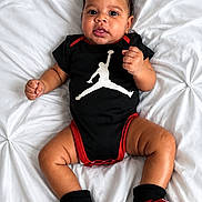 Jordan is registered to the contest to win money with this photo: baby, infant, onesie, black_clothing, red_clothing, socks, red_socks, white_blanket, textured_blanket, curly_hair, cute, lying_down, indoor, portrait, child, person, smiling, looking_at_camera, small_hands, adorable