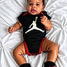 baby, infant, onesie, black_clothing, red_clothing, socks, red_socks, white_blanket, textured_blanket, curly_hair, cute, lying_down, indoor, portrait, child, person, smiling, looking_at_camera, small_hands, adorable