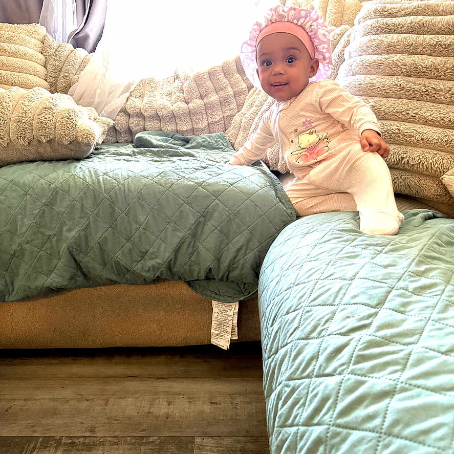 Avila is registered to the contest to win money with this photo: baby, child, infant, crown, smiling, sitting, sofa, couch, quilt, blanket, cushion, living_room, indoors, window, curtains, wooden_floor, clothing, portrait, home, cozy