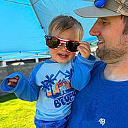 Camden is registered to the contest to win money with this photo: toddler, child, man, sunglasses, cap, blue_shirt, outdoor, sunny, canopy, grass, smile, hand, face, clothing, holding, portrait, daylight, hat, happy, family