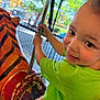 child, toddler, carousel, amusement_park, tiger, ride, green_shirt, plaid_shorts, pole, smiling, outdoor, daylight, playful, face, portrait, fun, rustic, vintage, metal, fence