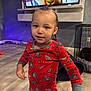 child, toddler, pajamas, dinosaur_print, indoor, floor, tv, cartoon, person, face, hair, hand, clothing, room, furniture, pet_crate, toy, curious, standing, young_child