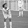 child, soccer, sports, field, uniform, goalpost, flag, grass, outdoor, team, athlete, game, sporting_equipment, young_player, white_clothing, knees_bent, standing, black_and_white, focus, action
