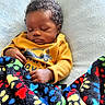 baby, sleeping, blanket, yellow_sweater, curly_hair, indoor, cushion, cozy, child, peaceful, resting, soft_texture, colorful, patterned_blanket, portrait, infant, hand, face, closeup, relaxing