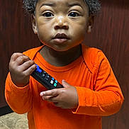 Righteous Parker is registered to the contest to win money with this photo: child, toddler, orange_shirt, remote_control, indoor, curious, person, portrait, brown_skin, short_hair, standing, wood_panel, tile_floor, clothing, hand, face, eyes, expression, cute, young