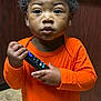 child, toddler, orange_shirt, remote_control, indoor, curious, person, portrait, brown_skin, short_hair, standing, wood_panel, tile_floor, clothing, hand, face, eyes, expression, cute, young