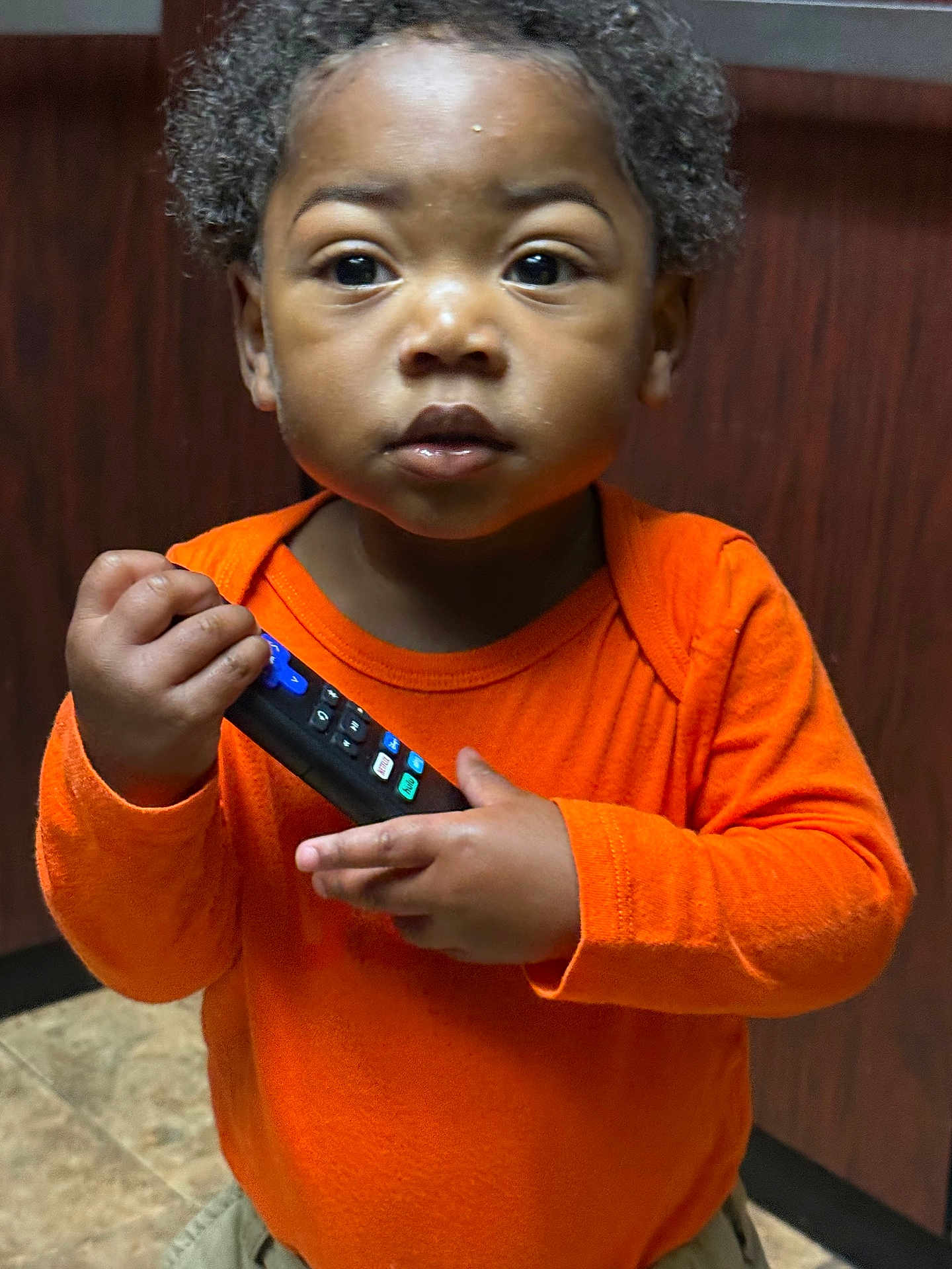 Righteous Parker is registered to the contest to win money with this photo: child, toddler, orange_shirt, remote_control, indoor, curious, person, portrait, brown_skin, short_hair, standing, wood_panel, tile_floor, clothing, hand, face, eyes, expression, cute, young