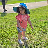 Camille is registered to the contest to win money with this photo: toddler, child, grass, outdoor, sunhat, pink_shirt, shorts, zebra_print, shoes, shadow, daylight, person, curly_hair, playground, blurred_background, adult, greenery, park, casual_clothing, summer