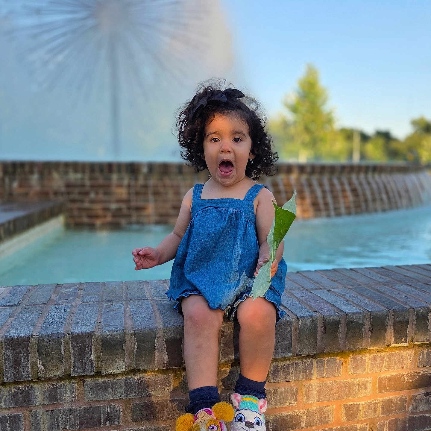 Camille joined the competition — help win amazing prizes! child, girl, blue_dress, brick_wall, fountain, outdoor, leaf, curly_hair, expression, sitting, footwear, slippers, water, daylight, nature, tree, sky, playful, surprised, sculpture