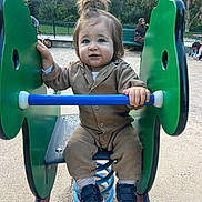 Jarod participe au concours pour gagner de l'argent avec cette photo : toddler, child, playground, spring_rocker, green, blue, brown_clothing, shoes, hair_tied_up, outdoor, sand, park, fence, bench, person, young_child, curious_expression, daylight, casual_wear, fun