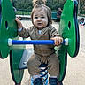 toddler, child, playground, spring_rocker, green, blue, brown_clothing, shoes, hair_tied_up, outdoor, sand, park, fence, bench, person, young_child, curious_expression, daylight, casual_wear, fun