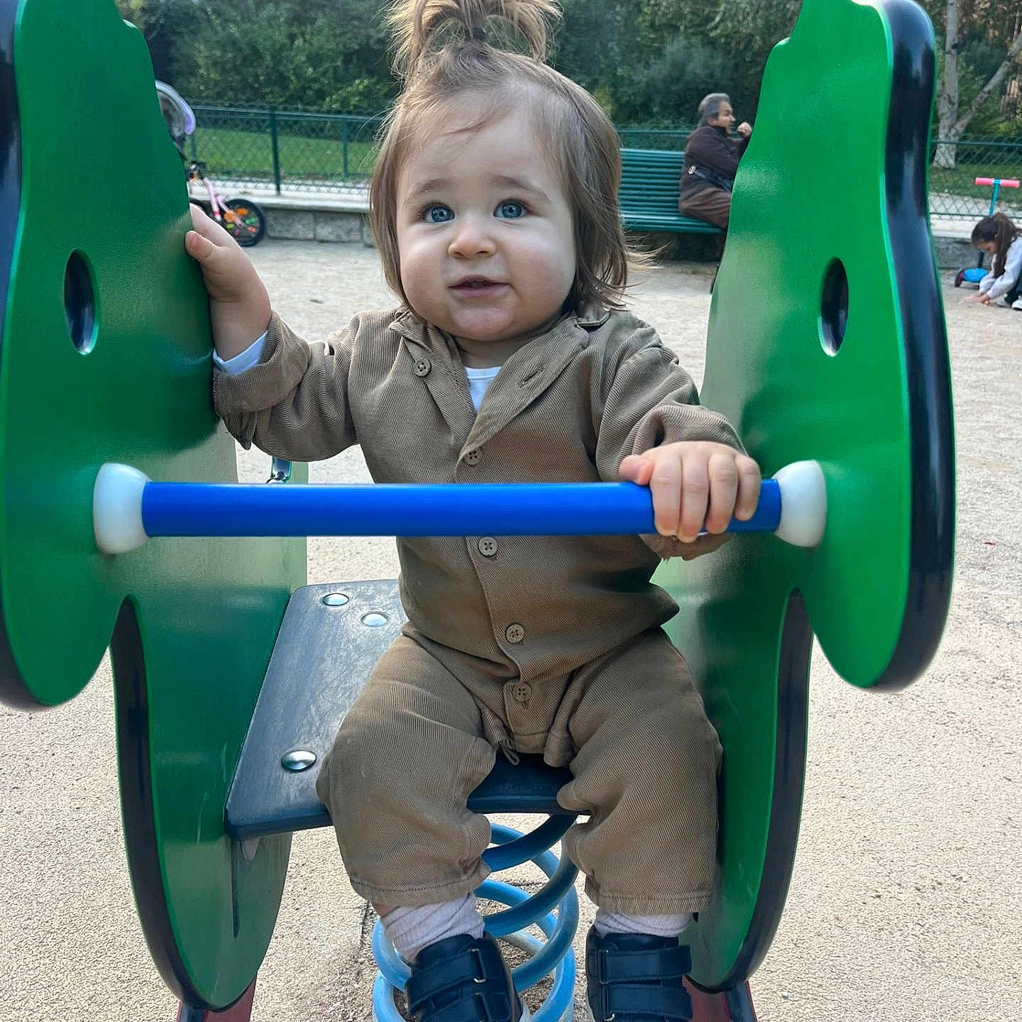 Jarod participe au concours pour gagner de l'argent avec cette photo : toddler, child, playground, spring_rocker, green, blue, brown_clothing, shoes, hair_tied_up, outdoor, sand, park, fence, bench, person, young_child, curious_expression, daylight, casual_wear, fun