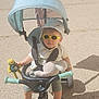 Lisandru participe au concours pour gagner de l'argent avec cette photo : toddler, child, sunglasses, hat, tricycle, stroller, toy, plush_toy, frog_doll, outdoor, sunny, street, summer_clothing, footwearless, shadow, handlebars, seatbelt, pavement, casual, cute