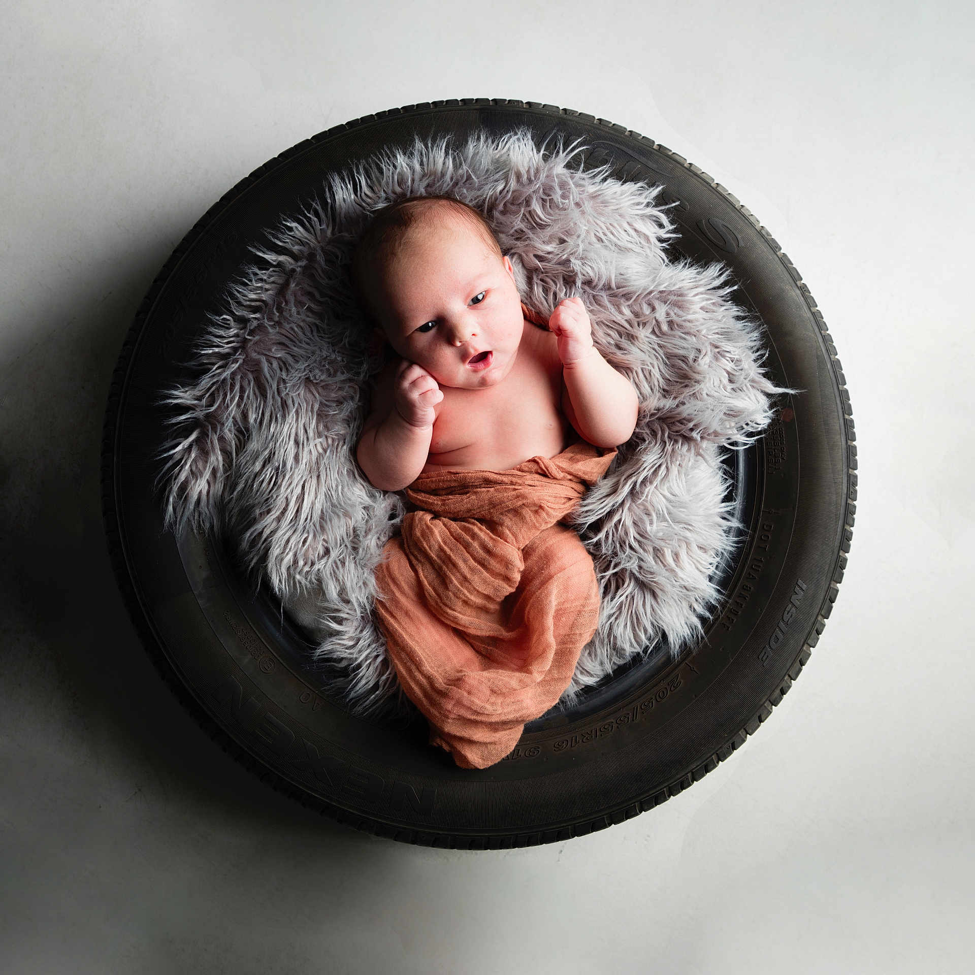 Leon is registered to the contest to win money with this photo: baby, infant, tire, blanket, fur, soft_texture, wrapped, curious, raised_fists, studio_lighting, floor, round, black, gray, orange_cloth, newborn, portrait, cute, indoor, minimalist