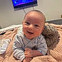 Leon is registered to the contest to win money with this photo: baby, smiling, blanket, plush_toy, indoor, television, child, happy, tummy_time, cozy, cute, person, soft_lighting, bedroom, infant, clothing, relaxed, home, comfort, young_child