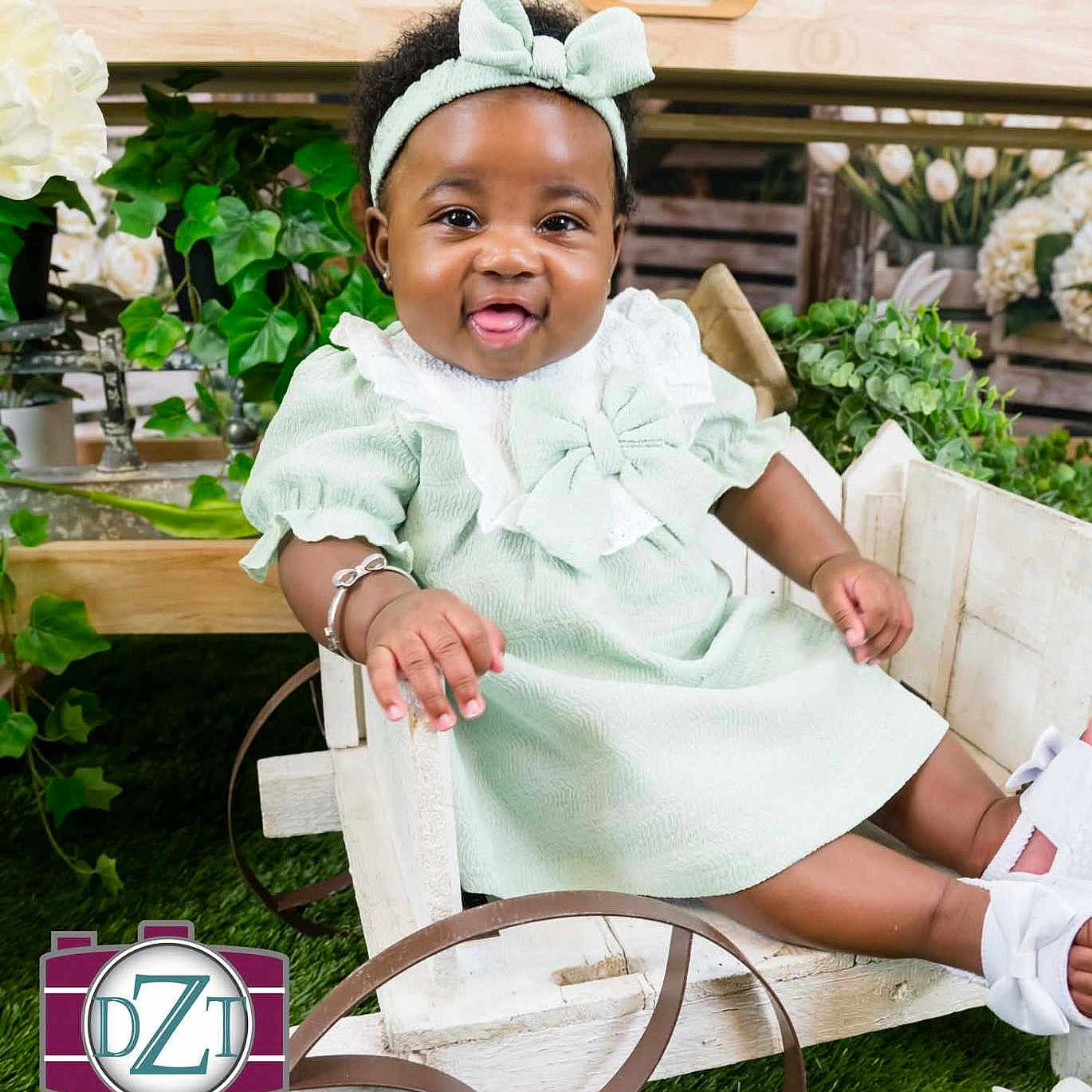 Amenna is registered to the contest to win money with this photo: baby, child, girl, smiling, headband, dress, bow, wooden_cart, greenery, plants, flowers, grass, bracelet, happy, sitting, portrait, footwear, white_shoes, indoor, photography_studio