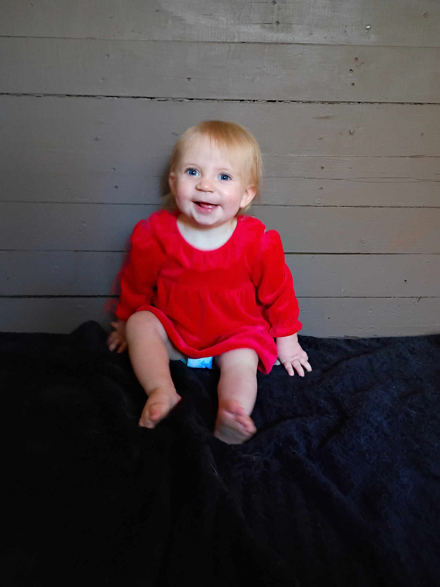 Brynleigh joined the competition — help win amazing prizes! baby, child, smiling, red_dress, sitting, blanket, wooden_wall, happy, indoors, person, cute, portrait, blue_eyes, blond_hair, infant, feet, hand, clothing, face, skin