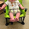 animal_shirt, baby, background, casual_clothing, child, concrete_floor, curious_expression, face, floor, footwear, green_chair, hands, indoor, infant, legs, person_walking, plaid_shorts, portable_chair, seated, young_child