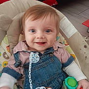 Kenzo participe au concours pour gagner de l'argent avec cette photo : baby, infant, smiling, blue_eyes, denim_overalls, striped_shirt, pacifier, toy, baby_seat, cushion, indoor, floor, red_blanket, child, happy, person, clothing, cute, young, portrait