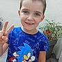 child, boy, peace_sign, blue_shirt, sonic, outdoor, plants, smile, short_hair, concrete_floor, casual_clothing, young, portrait, gesture, happy, front_view, daylight, pot, greenery, wall