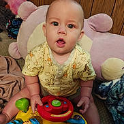 Wiliam is registered to the contest to win money with this photo: baby, child, toy, steering_wheel, plush, cushion, carpet, wood_paneling, yellow_clothing, sitting, indoor, hands, face, eyes, mouth_open, furniture, colorful, playtime, cute, infant